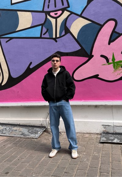 Sudadera negra con cremallera, camiseta blanca, pantalones vaqueros holgados de color azul claro y zapatillas blancas. El fondo presenta un colorido mural con tonos morados y rosas.