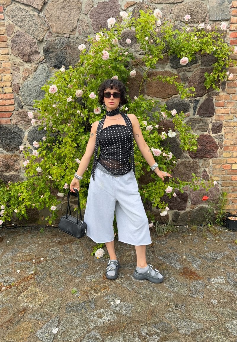 Personne portant un haut noir à pois, un pantalon large blanc, des sandales grises et des lunettes de soleil, debout devant un mur en pierre avec un buisson vert en fleurs.