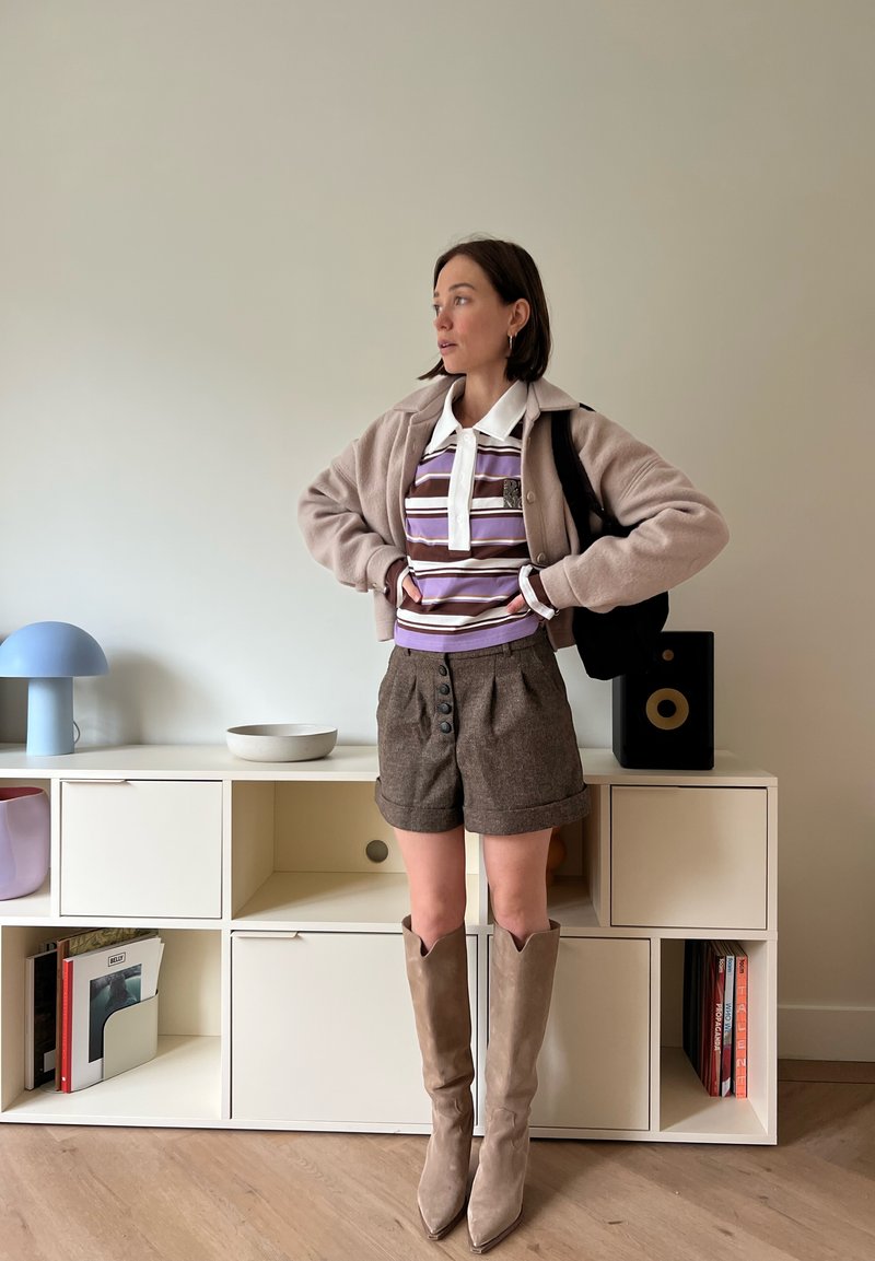 Femme aux cheveux courts et foncés portant un polo rayé, une veste beige, un short marron et des bottes beige montantes, debout les mains sur les hanches dans une pièce minimaliste.
