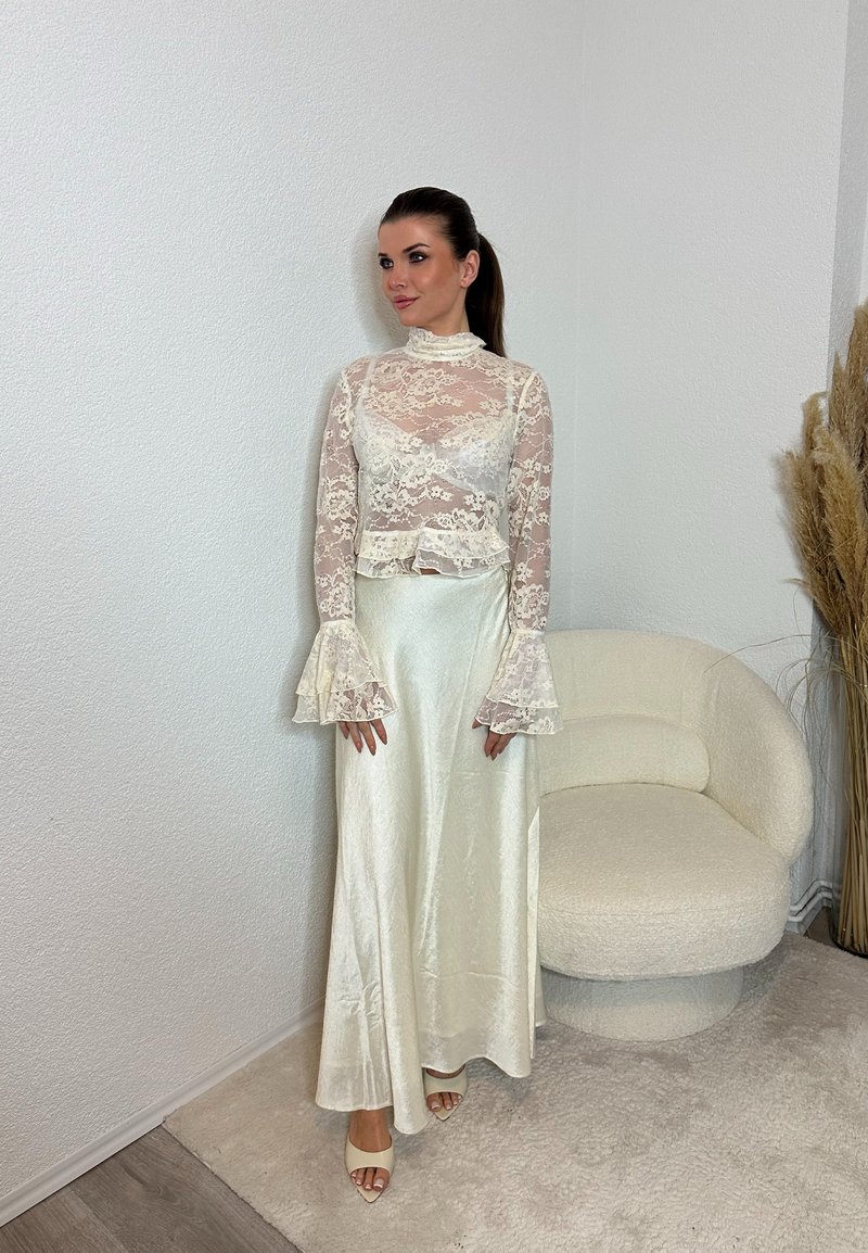 Γυναίκα στέκεται κοντά σε κρεμ καρέκλα, φοράει λευκό διαφανές δαντελένιο τοπ με καμπάνα μανίκια και μακριή λευκή σατέν φούστα, τα μαλλιά της σε αλογοουρά, ουδέτερο φόντο.