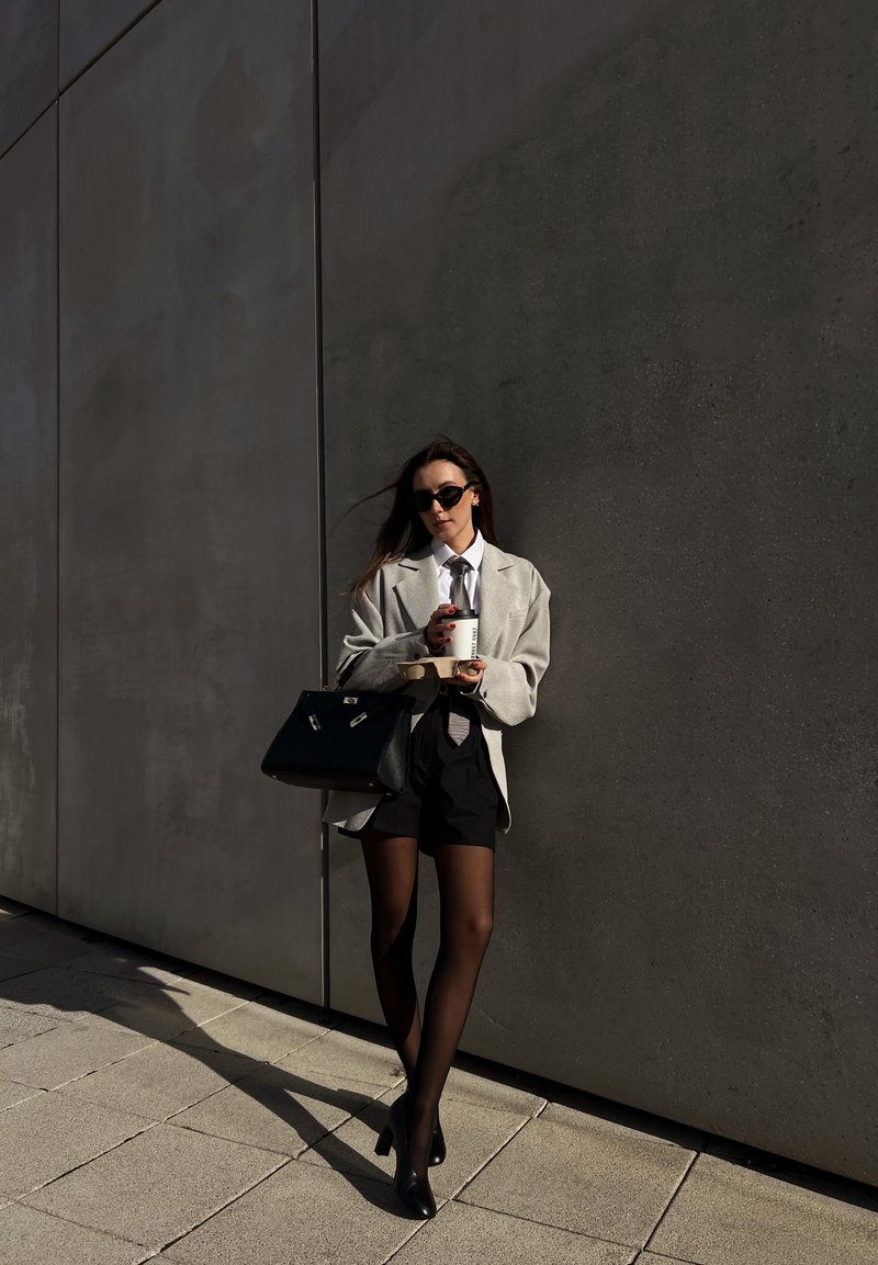 Blazer gris oversize sur une chemise blanche, short noir, collants noirs transparents et chaussures à talons hauts. Sac à main noir, tenant un verre.