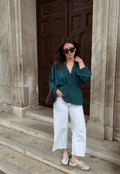 Mujer con blusa azul verdosa, pantalones blancos cortos, zapatillas deportivas y gafas de sol, de pie sobre escalones de piedra frente a una gran puerta de madera.