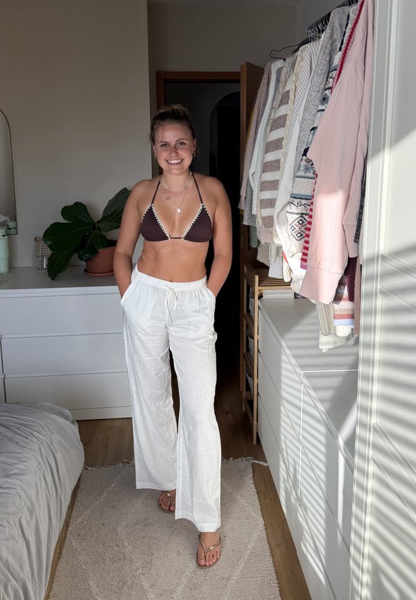 Młoda kobieta w brązowym bikini i białych spodniach stoi uśmiechnięta w sypialni z wiszącymi ubraniami i rośliną doniczkową w pobliżu.