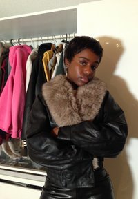 Jeune personne aux cheveux courts portant une veste en cuir noire avec un grand col en fourrure, debout, les bras croisés devant une armoire.