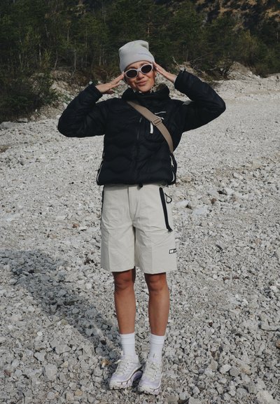 Persona con chaqueta negra, pantalones cortos beige, zapatillas blancas, gorro y gafas de sol, de pie en un terreno rocoso con árboles al fondo.