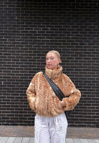 Casaco de pelagem sintética em castanho claro com manchas brancas, gola alta e mangas oversized. Usado com calças cargo brancas e uma mala de ombro preta com tachas.