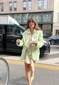 Manteau en mohair vert menthe, robe crème à volants, chaussures claires et accessoires. Scène urbaine avec des véhicules et des bâtiments.