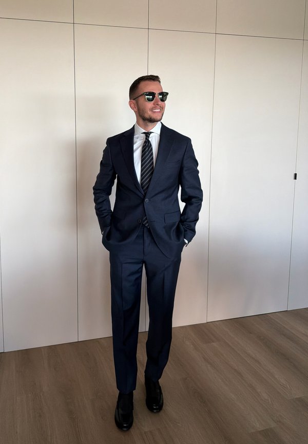 Hombre vestido con un traje azul oscuro, corbata a rayas, zapatos negros y gafas de sol, de pie en el interior contra una pared clara con las manos en los bolsillos.