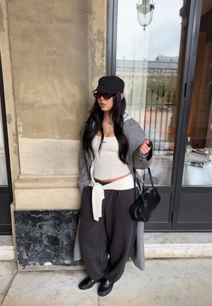 Mujer con cabello largo y negro, que lleva una gorra negra, gafas de sol, camiseta blanca, abrigo gris, pantalones negros y anchos, y sostiene un bolso negro, de pie junto a un edificio.