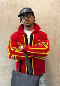 Άνδρας με μαύρο καπέλο, γυαλιά, κόκκινο πουλόβερ με σχέδια και ρολόι χειρός, κρατώντας μαύρη δερμάτινη τσάντα ώμου μπροστά σε έναν απλό τσιμεντένιο τοίχο.