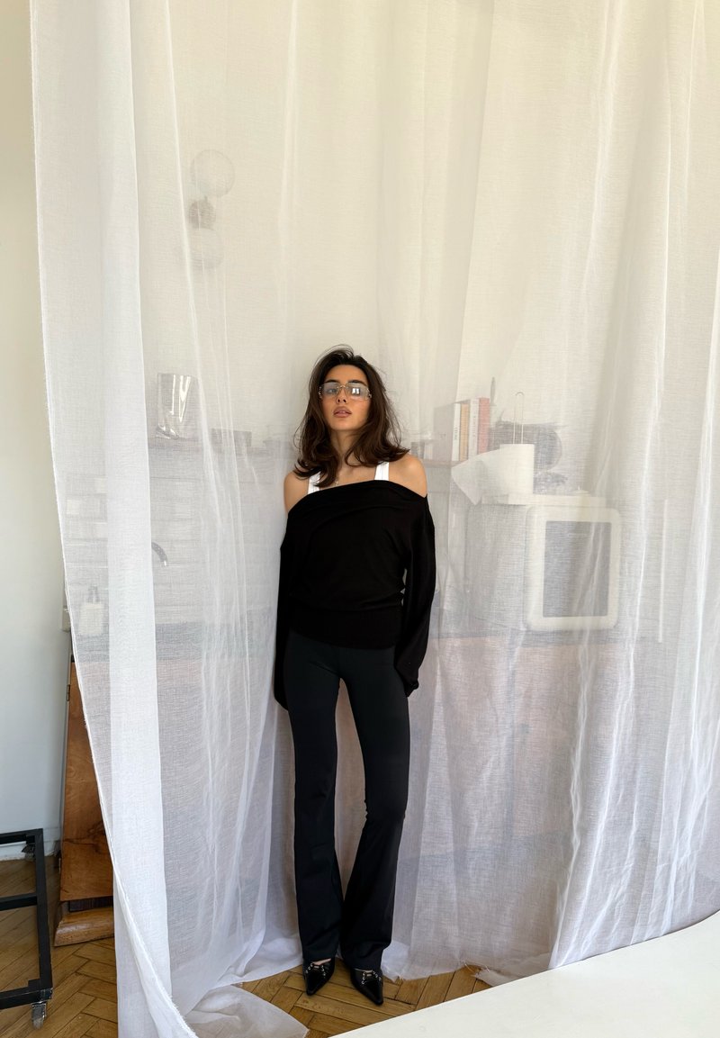 Ženska z dolgimi lasmi, ki nosi očala, črna majica brez ramen in črne hlače, stoji za prosojnimi belimi zavesami v sodobni sobi.