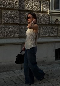 Γυναίκα με πουά αμάνικο τοπ και τζιν με φαρδύ πόδι περπατά δίπλα σε υφασμένη πέτρινη τοίχο, κρατώντας μια μαύρη τσάντα και κουβαλώντας τα μαλλιά της.