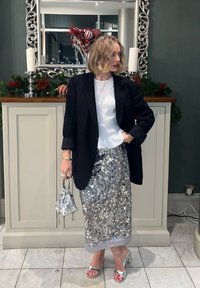 Jupe argentée à sequins avec un haut blanc texturé et un blazer noir. Tenant un petit sac argenté, debout sur un sol carrelé blanc, avec une décoration festive.