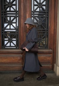 Une femme en manteau gris, chapeau assorti et chaussures foncées passe devant une porte en bois ornée en portant un grand sac à main marron foncé.
