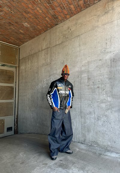 Hombre con vaqueros oscuros de gran tamaño, chaqueta de carreras negra con logotipos, gorro de rayas, zapatos slip-on a cuadros y gafas de sol, de pie junto a una pared de hormigón.