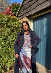 Lang marineblå frakk over et marmorert skjørt med blå, hvite og burgunder striper, paret med en stripete polo skjorte. Grønt bladverk og blå dør i bakgrunnen.