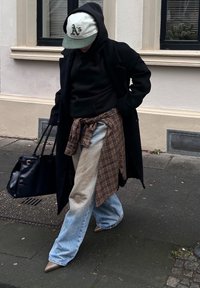 Schwarzer langer Mantel über einem schwarzen Kapuzenpullover, karierte Hemd um die Taille gebunden, hellblaue Jeans und beige spitze Schuhe. Schwarze Handtasche in der Hand.