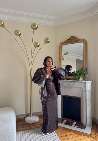 Vestido de satén marrón con estampado floral, chaqueta oscura de gran tamaño, calzado plateado, de pie junto a una chimenea de mármol blanco y una lámpara de pie dorada.