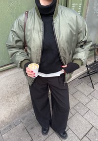 Personne portant une veste bomber verte et un col roulé noir tient un latte avec une mousse en forme de cœur, debout sur un trottoir en ville.