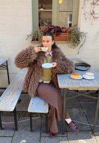 Cappotto marrone soffice, maglione verde, pantaloni marroni a gamba larga. Tiene in mano una tazza da tè, seduto a un tavolo con un dolce e un caffè in piatti bianchi.