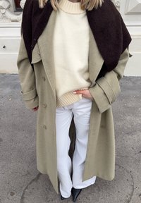 Femme portant un long manteau beige, un pull crème, un pantalon blanc, des chaussures noires à bouts pointus, et un châle brun foncé drapé sur les épaules, debout sur le trottoir.