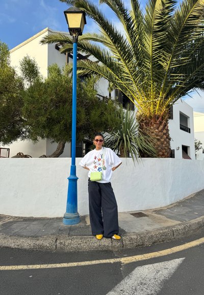 Camiseta blanca oversize con patrones coloridos, pantalones de pierna ancha a rayas negras, zapatos amarillos y un bolso cruzado verde. Farola azul al fondo.