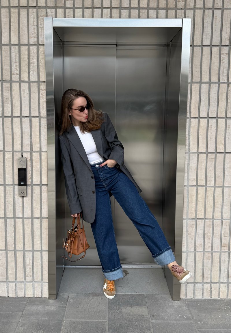 Frau mit Sonnenbrille, grauem Blazer, weißem Hemd und hochgekrempelten Jeans lehnt spielerisch an einem metallischen Aufzugstürrahmen und hält eine braune Handtasche.