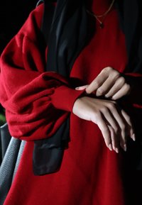 Piros pulóver sokféle ujjakkal, texturált anyagból, szűk mandzsettával. Fekete sál a vállakon; a kezek laza helyzetben.