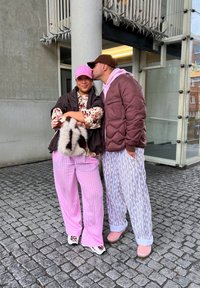 Dva lidé stojí venku. Jeden má na sobě květinovou blůzu, hnědou vestu, růžové pruhované kalhoty a koženou kabelku. Druhý má na sobě hnědou prošívanou bundu a vzorované kalhoty.