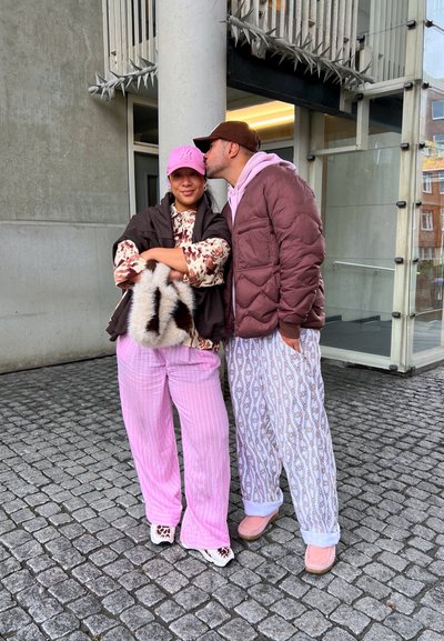 Dos personas están de pie al aire libre. Una de ellas lleva una blusa floral, un chaleco marrón, pantalones a rayas rosas y un bolso de piel de animal. La otra lleva una chaqueta de abrigo marrón y pantalones con un patrón.