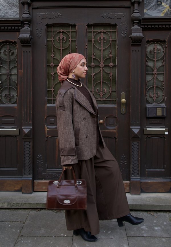 Vrouw met een bruine blazer, wijde broek, roze hoofddoek met patroon, houdend een leren handtas, lopend langs sierlijke donkere houten deuren.