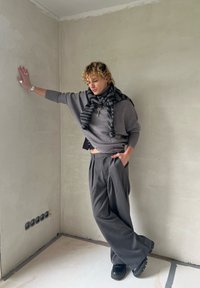Pull gris surdimensionné, écharpe rayée noir et gris, pantalon plissé taille haute et chaussures noires ; posé contre un mur uni.
