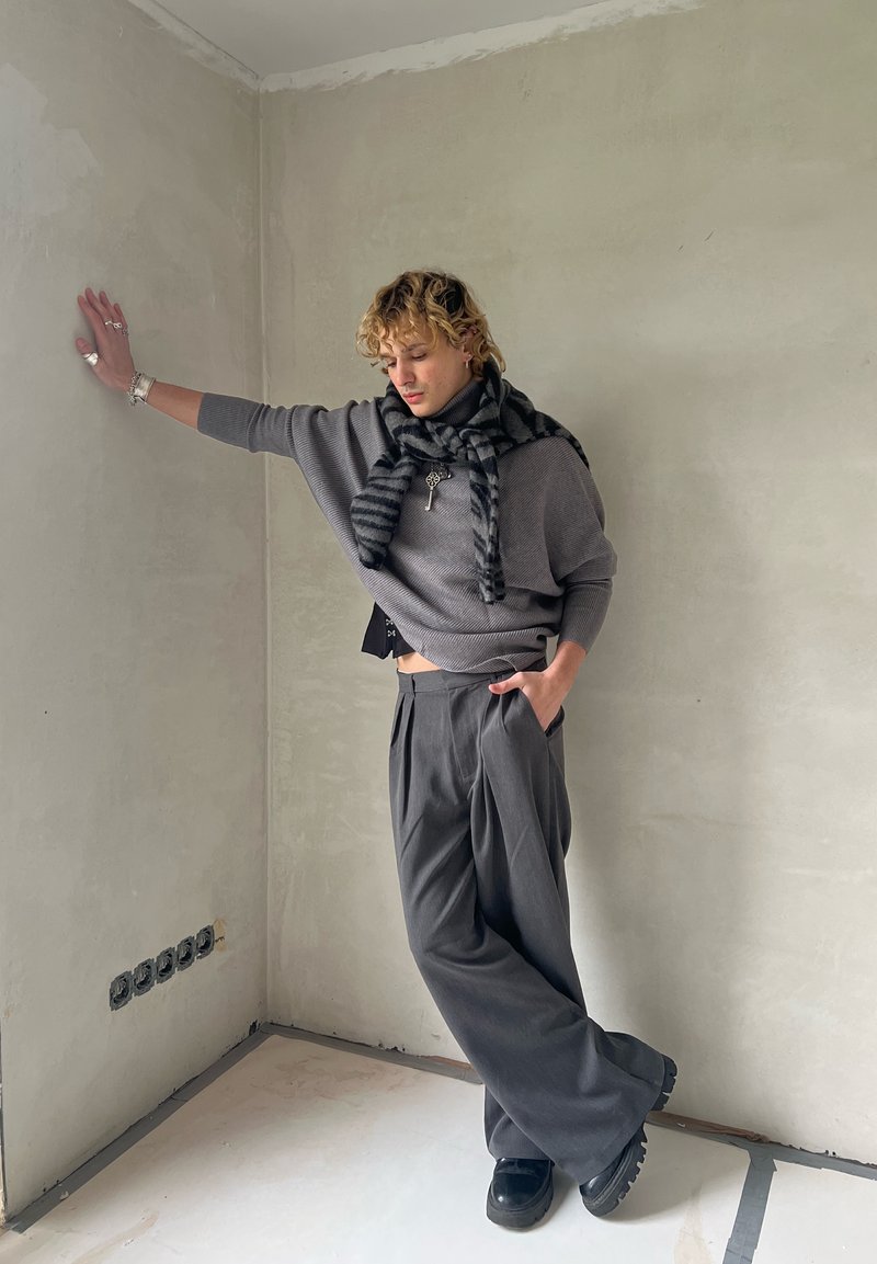 Pull gris surdimensionné, écharpe rayée noir et gris, pantalon plissé taille haute et chaussures noires ; posé contre un mur uni.