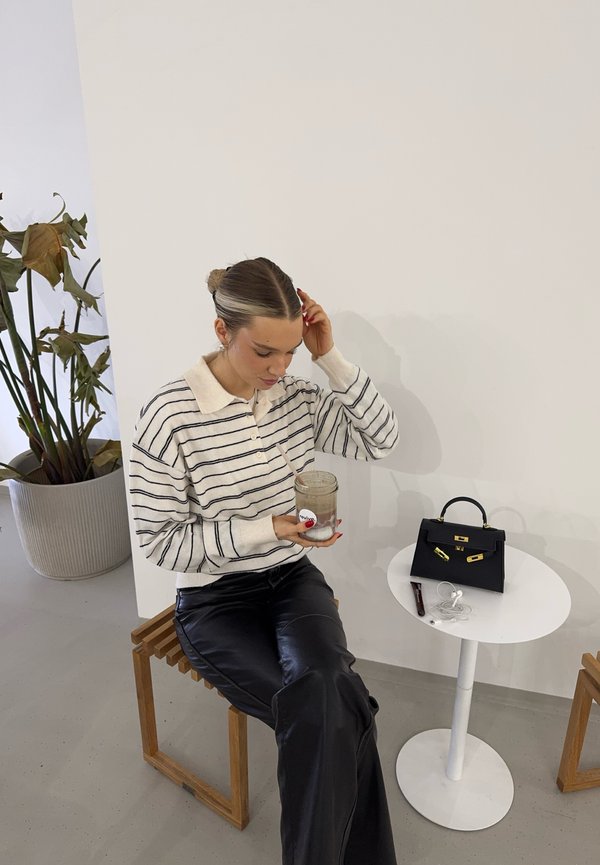 Csíkos pulóver bézs gallérral, fekete bőrnadrág, kezében egy üveg. Mellette egy kis fekete kézitáska egy fehér asztalon. Háttérben barna virágtartó.
