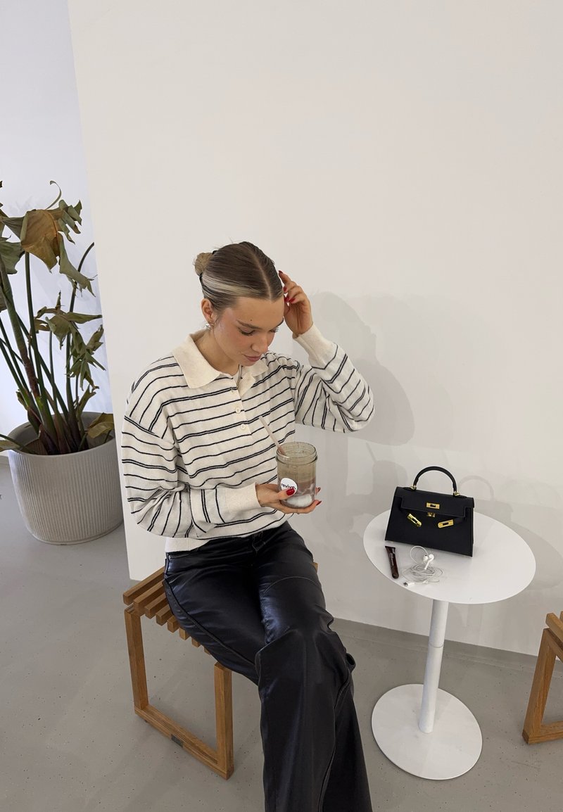 Pull rayé avec un col beige, pantalon en cuir noir, tenant un bocal. Un petit sac à main noir sur une table blanche à côté d'elle. Planteur marron en arrière-plan.