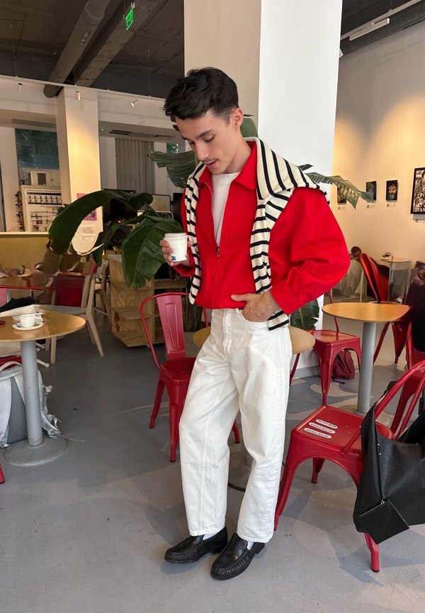 Jeune homme en veste rouge et pantalon blanc tenant une tasse dans un café avec des chaises rouges, des tables rondes, une grande plante verte et des œuvres murales.