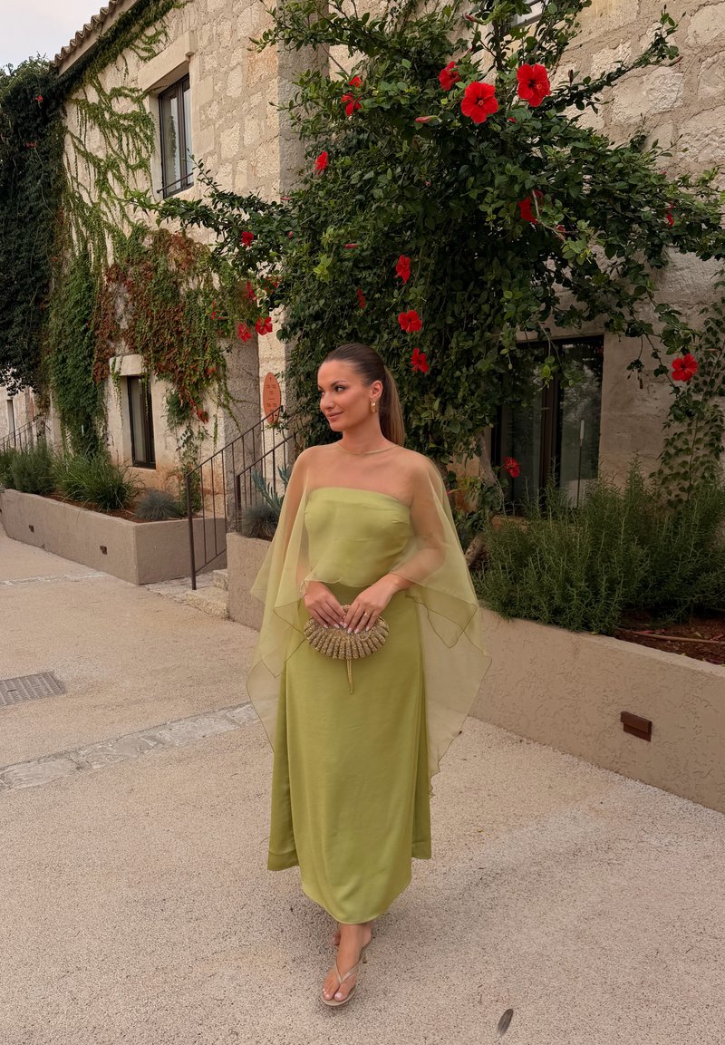 Femme en robe bustier vert clair et châle transparent tenant une pochette tissée, debout dans une rue pavée avec des bâtiments en pierre et des fleurs rouges à proximité.