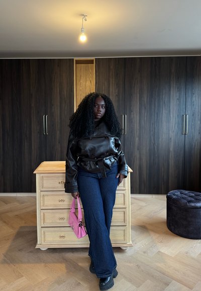 Persona con cabello rizado, chaqueta de cuero negra, pantalones vaqueros azules y botas negras, de pie en el interior sosteniendo un bolso rosa cerca de una cómoda de madera.