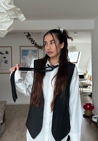 Gilet noir porté sur une chemise blanche, avec une texture de tissu lisse. Le gilet comporte des boutons et est assorti d'une cravate noire tenue à la main.