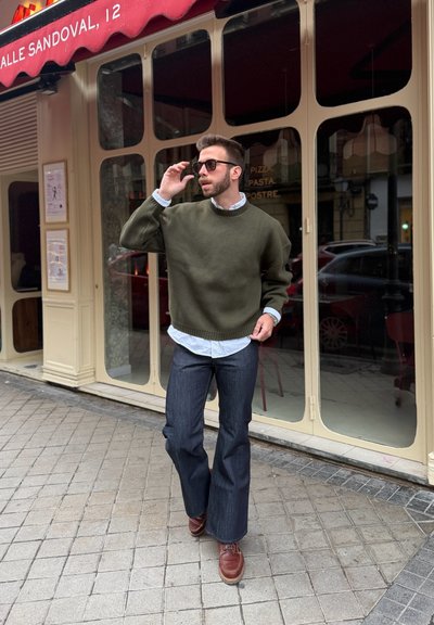 Hombre con gafas de sol, suéter verde, camisa blanca y vaqueros acampanados caminando frente a un restaurante con marcos de ventana beige y un toldo rojo.