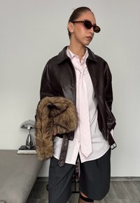 Veste bomber en cuir marron sur une chemise rayée rose, assortie à un short gris, tenant un vêtement orné de fourrure. Des lunettes de soleil noires complètent le look.