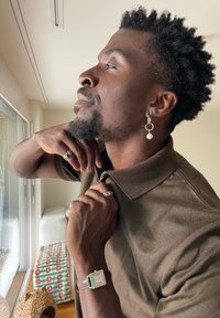 Muž s náušnicí a prsteny zapínající hnědé polo tričko, nosící hodinky se čtvercovým ciferníkem, stojící u okna v jasné místnosti.