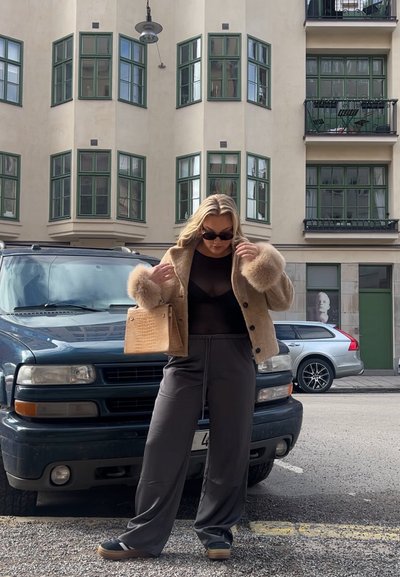 Chaqueta beige con borde de piel sintética, blusa negra, pantalones grises de pierna ancha y zapatos negros. Sosteniendo un bolso de patrón cocodrilo en color marrón. Fondo urbano.