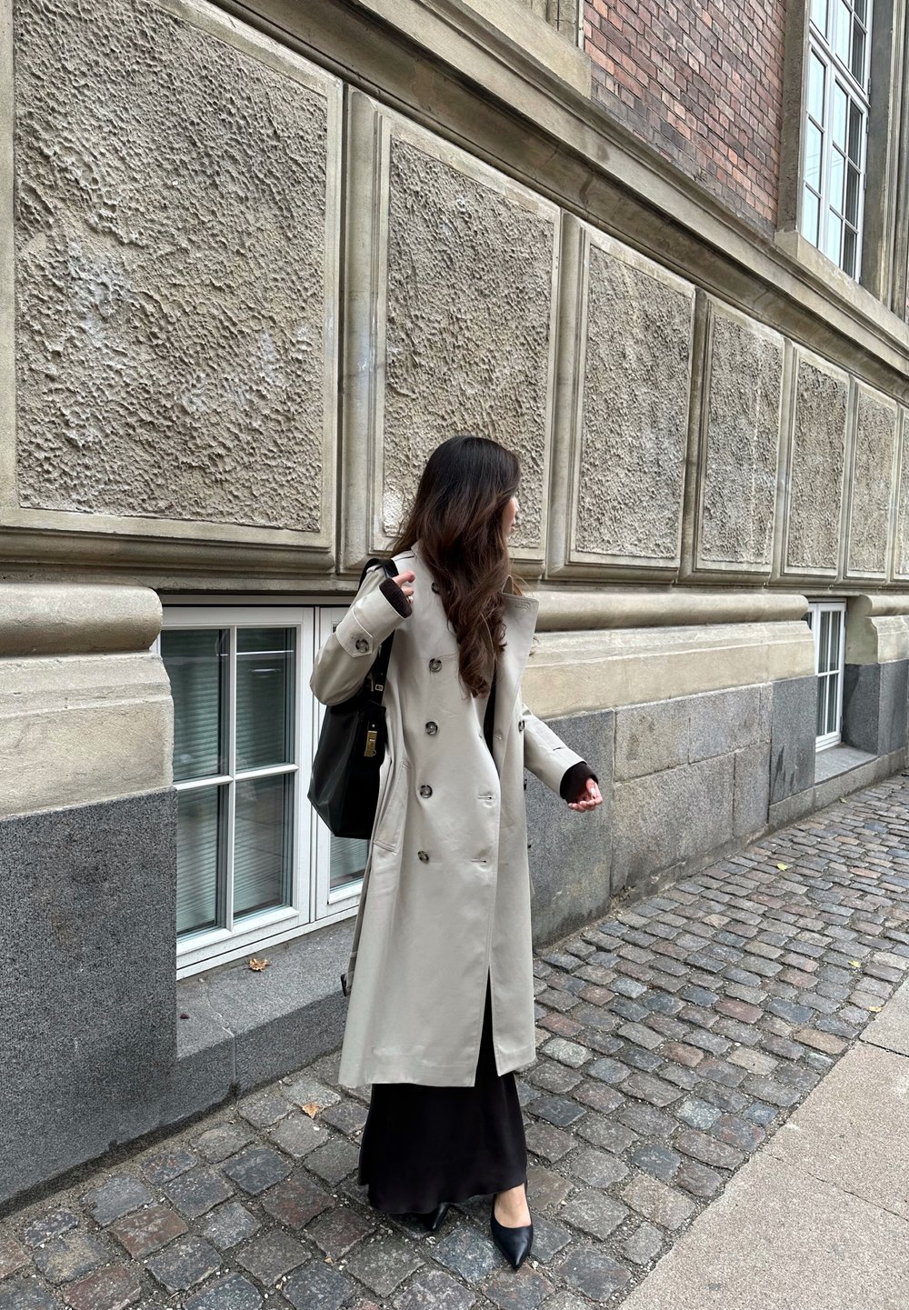 Μπεζ παλτό trench με εφαρμοστή γραμμή, διπλό κούμπωμα και δομημένο γιακά. Φορεμένο πάνω από ένα μαύρο φόρεμα maxi και σε συνδυασμό με μαύρες γόβες.