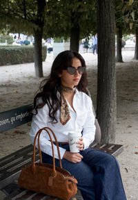 Sac à main en daim brun clair de forme allongée avec deux poignées ; modèle en chemise blanche et foulard à motifs tenant une tasse de café, assis sur un banc.