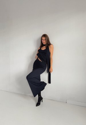 Vestido negro sin mangas con un diseño drapeado, cortes laterales y una silueta hasta el suelo, combinado con zapatos de tacón negros.