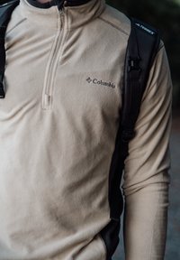 Beige fleece-huppari, jossa on puoliveto suunnittelu, jossa on Columbia-logo rinnan kohdalla ja mustat olkaimet lisätoimintoja varten.