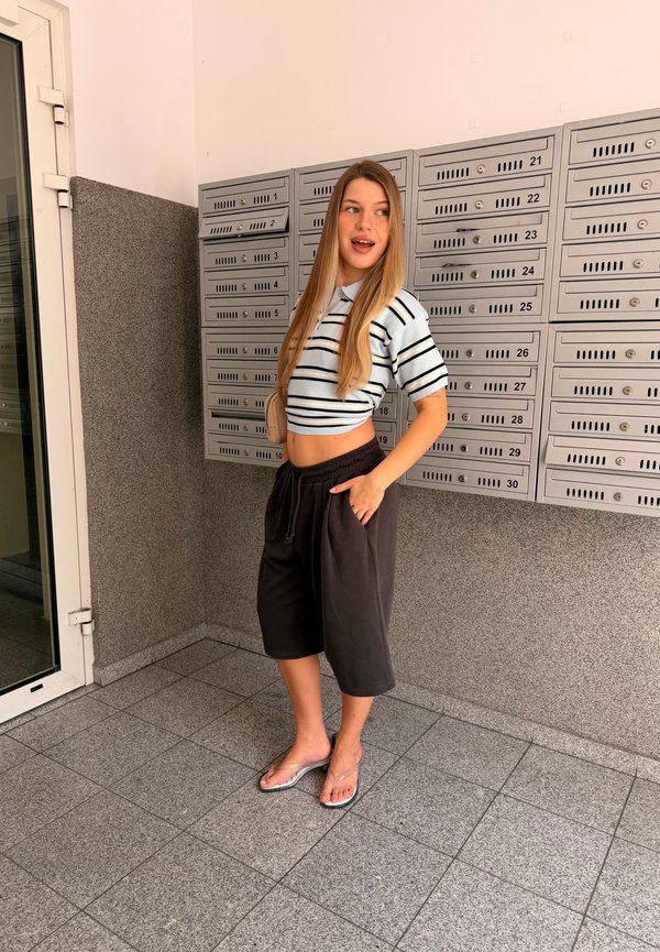 Jeune femme en crop top rayé et short debout près de rangées de boîtes aux lettres en métal dans un couloir carrelé, regardant sur le côté avec un sourire.