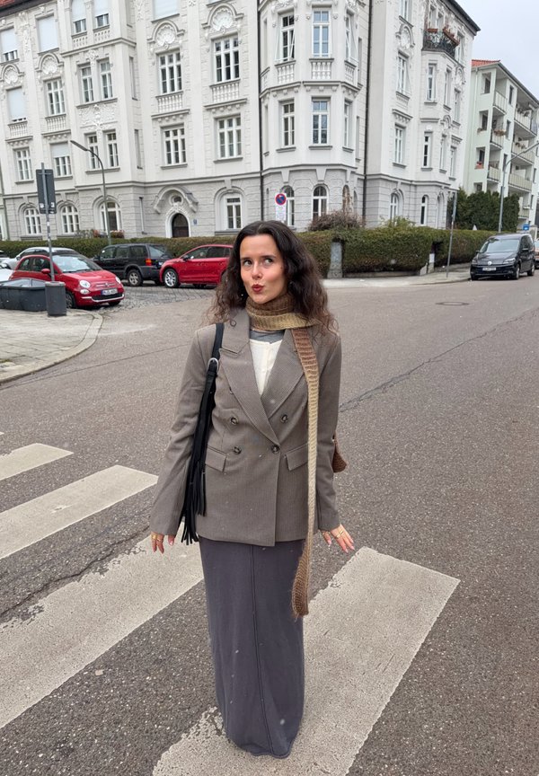 Grey and brown houndstooth double-breasted blazer, grey long skirt, knitted scarf, and a black fringed bag, standing on a pedestrian crossing.