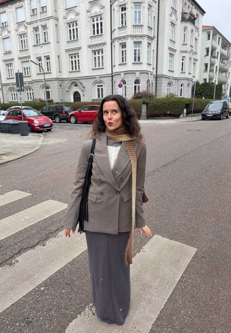 Grey and brown houndstooth double-breasted blazer, grey long skirt, knitted scarf, and a black fringed bag, standing on a pedestrian crossing.